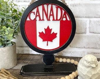 Puede incluir: Un letrero de madera de Canadá con una bandera canadiense roja y blanca y la palabra "CANADA" en letras rojas. El letrero está en un soporte negro con un borde de cuentas. El letrero está sobre una superficie de madera con una hebra de cuentas de madera y una pequeña figurita de pájaro blanco.