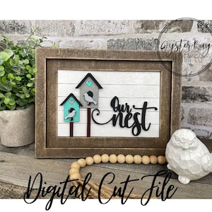 Pode incluir: Um design de arquivo de corte digital com duas casas de pássaros com pássaros, um coração e o texto "Our Nest". O design é emoldurado em uma moldura marrom rústica e exibido em uma superfície de madeira.
