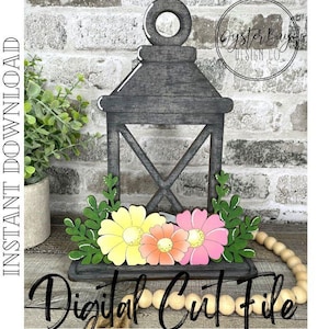 Puede incluir: Una linterna de madera gris con un diseño floral. La linterna tiene un diseño de flores blancas, rosas y amarillas con hojas verdes. La linterna está sentada sobre una superficie de madera con una cuerda de cuentas de madera.