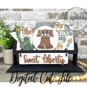 May include: A set of four wooden cut files featuring a Liberty Bell, a star with the Statue of Liberty, a sign that says "Sweet Liberty", and a sign that says "Let Freedom Ring" with fireworks. The cut files are on a black stand with a wooden bead garland.