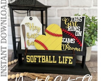 May include: A digital cut file for a softball themed tiered tray decor. The design includes a heart-shaped softball, a sign that says "This girl runs on seams & dreams", and a tag that says "Hey batter batter".