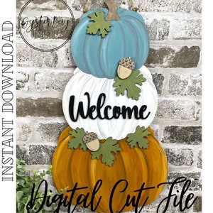 Puede incluir: Un colgador de puerta de calabaza en capas con una calabaza azul verdosa, blanca y naranja. La calabaza del medio tiene la palabra "Welcome" escrita en ella. Las calabazas están decoradas con hojas verdes y bellotas.