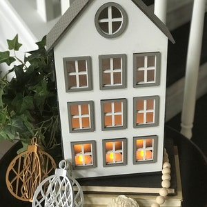May include: A white wooden house shaped candle holder with gray trim and nine windows. The windows have small lights inside. The house is sitting on a stack of books with a white beaded garland and two white and gold geometric ornaments.