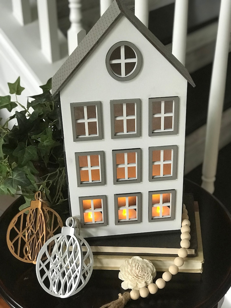 May include: A white wooden house shaped candle holder with gray trim and nine windows. The windows have small lights inside. The house is sitting on a stack of books with a white beaded garland and two white and gold geometric ornaments.