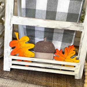 Könnte beinhalten: Holzdekorationen zum Thema Herbst, darunter zwei orangefarbene und gelbe Blätter und eine braune Eichel, werden auf einem weißen Regal mit einem schwarz-weiß karierten Handtuch dahinter präsentiert.