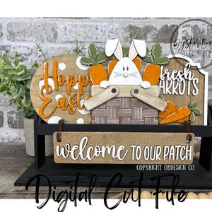 Op de afbeelding: Houten paasdecoratie met een konijn in een mand en wortels, de woorden "Hopp Easter" en "fresh carrots". Een bord hieronder luidt "welcome TO OUR PATCH". Het ontwerp staat op een zwarte standaard.