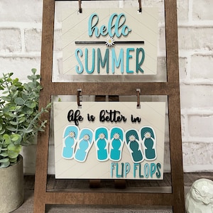 Könnte beinhalten: Holzschild mit zwei Tafeln. Die obere Tafel zeigt "hello SUMMER" in Blau und Weiß. Die untere Tafel zeigt "life is better in FLIP FLOPS" mit blauen Flip-Flop-Grafiken. Das Schild hat einen braunen Holzrahmen.