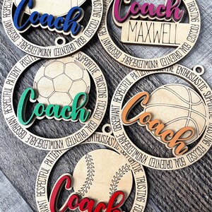Peut inclure: Cinq ornements en bois avec diff&eacute;rents motifs de ballons de sport. Chaque ornement a un cercle noir avec le mot "Coach" dans une couleur diff&eacute;rente et un ballon de sport diff&eacute;rent au centre. Les mots "GOAL ORIENTED, KNOWLEDGEABLE, OBSERVANT, RESPECTFUL, PATIENT, POSITIVE, ENTHUSIASTIC, SUPPORTIVE, TRUSTING, FOCUSED" sont &eacute;crits autour de l'ext&eacute;rieur de chaque cercle.