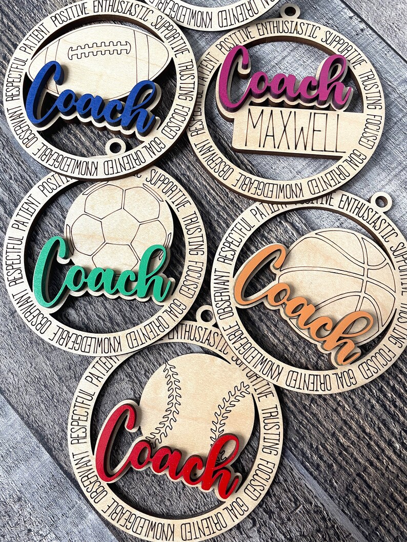 Peut inclure: Cinq ornements en bois avec diff&eacute;rents motifs de ballons de sport. Chaque ornement a un cercle noir avec le mot "Coach" dans une couleur diff&eacute;rente et un ballon de sport diff&eacute;rent au centre. Les mots "GOAL ORIENTED, KNOWLEDGEABLE, OBSERVANT, RESPECTFUL, PATIENT, POSITIVE, ENTHUSIASTIC, SUPPORTIVE, TRUSTING, FOCUSED" sont &eacute;crits autour de l'ext&eacute;rieur de chaque cercle.