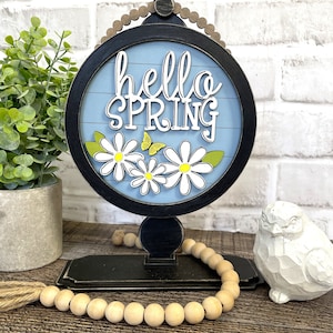 Peut inclure: Une pancarte en bois avec un fond bleu et le texte "hello spring" en blanc. La pancarte est décorée de marguerites blanches et de papillons jaunes. La pancarte est sur un support noir avec une guirlande de perles en bois.