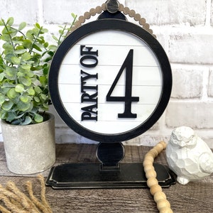 Könnte beinhalten: Schwarze und weiße Holztischnummer mit dem Text "Party of 4" auf weißem Hintergrund mit schwarzen Streifen. Die Nummer befindet sich auf einem schwarzen Ständer mit einer Perlenkrone.