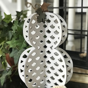 May include: White wooden pumpkin with a cut-out design. The pumpkin is layered and has a brown stem and leaf.