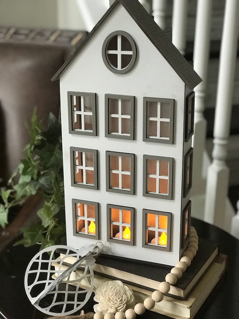 May include: A white wooden house-shaped candle holder with nine windows and a round window at the top. The windows have a brown frame and are lit with candles. The candle holder is sitting on a stack of books with a wooden bead garland.