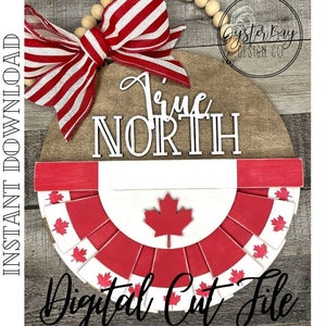 Op de afbeelding: Een rond houten bord met de woorden "True North" in witte letters. Het bord is versierd met een rood-wit gestreepte strik, een rood-witte Canadese vlag en een kralenkrans. Het bord is gelabeld met "Digital Cut File" in cursief.