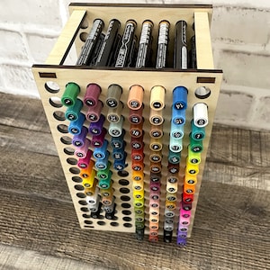 May include: A wooden stand with a grid of holes to hold markers. The stand is filled with colorful markers, with the numbers 1 through 80 visible on the caps. A white marker with the text "TOOLI-ART Acrylic Paint Pen" is lying on a wooden surface.