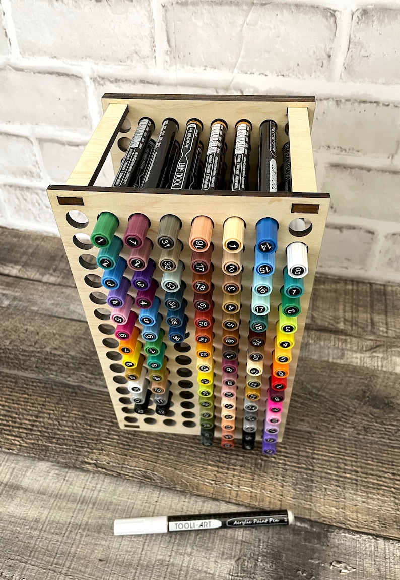 May include: A wooden stand with a grid of holes to hold markers. The stand is filled with colorful markers, with the numbers 1 through 80 visible on the caps. A white marker with the text "TOOLI-ART Acrylic Paint Pen" is lying on a wooden surface.