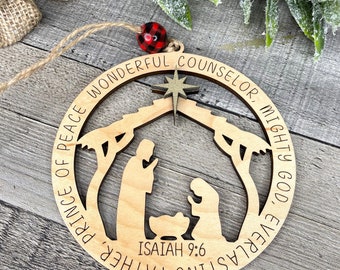 May include: A wooden Christmas bauble featuring a cut-out nativity scene. The bauble is round and inscribed with the words "Everlasting Father, Prince of Peace, Wonderful Counselor, Mighty God" and "Isaiah 9:6" around the edge.