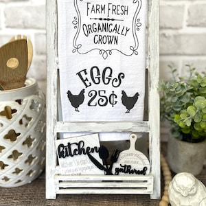 May include: White wooden stand with a white towel that says "Farm Fresh Organically Grown Eggs 25¢". There are two black chicken silhouettes on the towel. The stand has a shelf with a black wooden sign that says "kitchen" and another black wooden sign that says "The fondest memories are made gathered".