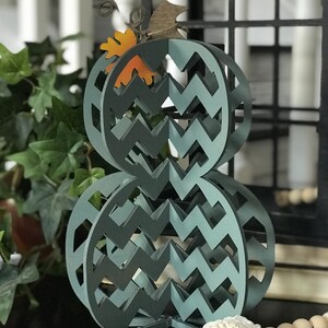 May include: A teal blue wooden pumpkin with a chevron pattern. The pumpkin is made of multiple layers and has a small orange leaf on top.