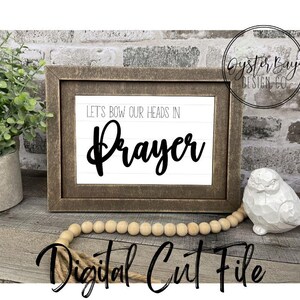 Puede incluir: Un letrero de madera marrón con marco de madera, fondo blanco y texto negro que dice "Let's bow our heads in Prayer". El letrero está colocado sobre una superficie de madera con una cadena de cuentas de madera y una figurita de pájaro blanca.