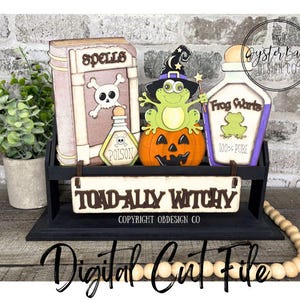 Könnte beinhalten: Ein Halloween-Themen-Holzregal mit dekorativen Ausschnitten. Enthält ein Buch mit der Aufschrift "Spells", eine Giftflasche, einen Frosch auf einem Kürbis und eine "Frog Warts"-Flasche. Das Schild lautet "TOAD-ALLY WITCHY".