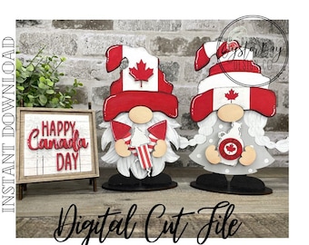 May include: Two wooden gnome figures decorated for Canada Day. The gnomes are wearing red and white hats with a Canadian flag design. One gnome is holding a red and white candy cane and the other is holding a bottle of maple syrup. The sign says "Happy Canada Day".
