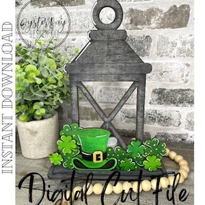Puede incluir: Una linterna de madera con un sombrero de duende verde y hojas de trébol. La linterna está pintada en un acabado gris desgastado. El texto "Digital Cut File" es visible en la parte inferior de la imagen.
