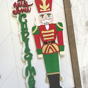 May include: A wooden nutcracker figurine with a red and gold uniform, green pants, and a black hat. The figurine is standing next to a wooden sign that says "nuts about Christmas" in green and red letters.
