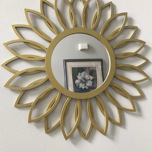 May include: A gold-colored, circular mirror with a sunflower design. The mirror has a round, reflective surface and is surrounded by a frame of overlapping petals.
