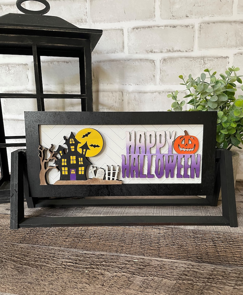 May include: A black wooden Halloween decoration with a haunted house, bats, and a pumpkin. The words "HAPPY HALLOWEEN" are displayed in purple and white. The decoration is on a wooden surface.