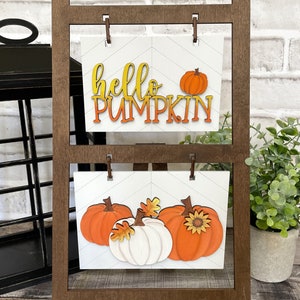 K&ouml;nnte beinhalten: Ein Holzst&auml;nder mit zwei wei&szlig;en Schildern mit Herbstdekoration. Das obere Schild sagt "hello pumpkin" in orangefarbenen und gelben Buchstaben. Das untere Schild zeigt drei K&uuml;rbisse, einen wei&szlig;en und zwei orangefarbene, mit Herbstbl&auml;ttern und einer Sonnenblume.