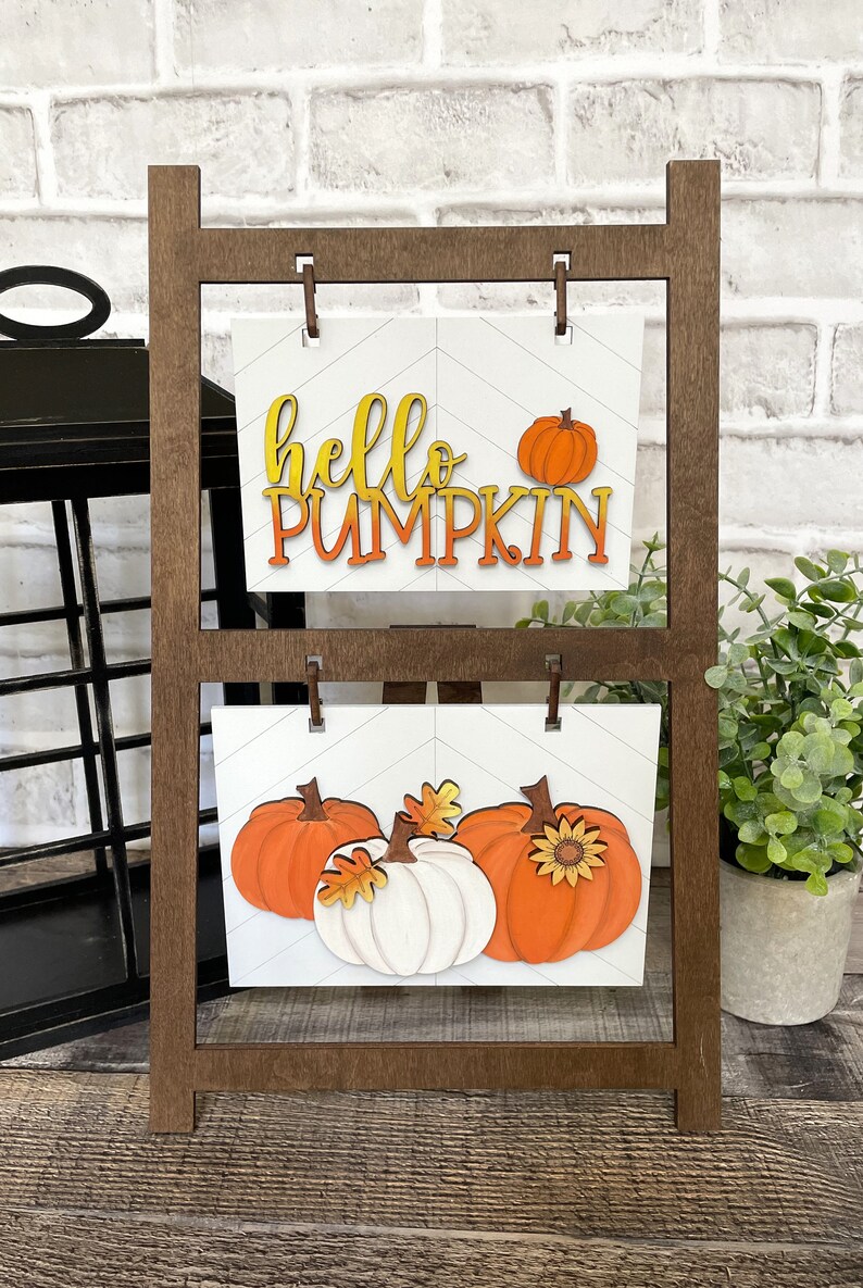 K&ouml;nnte beinhalten: Ein Holzst&auml;nder mit zwei wei&szlig;en Schildern mit Herbstdekoration. Das obere Schild sagt "hello pumpkin" in orangefarbenen und gelben Buchstaben. Das untere Schild zeigt drei K&uuml;rbisse, einen wei&szlig;en und zwei orangefarbene, mit Herbstbl&auml;ttern und einer Sonnenblume.