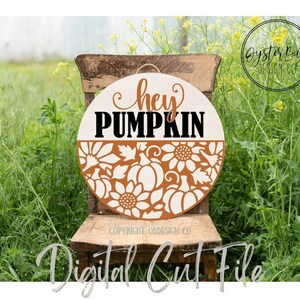 K&ouml;nnte beinhalten: Runder Holzschild mit den Worten "hey PUMPKIN" in Braun und Schwarz. Die untere H&auml;lfte zeigt ein ausgeschnittenes Design aus K&uuml;rbissen und Sonnenblumen in Hellbraun. Das Schild h&auml;ngt an einem Holzstuhl in einem Feld.