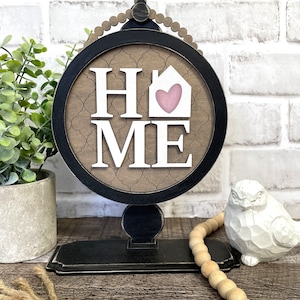 May include: A black and brown wooden sign with the word "HOME" in white letters. The "O" in home is a house shape with a pink heart inside. The sign is on a black stand with a beaded trim.