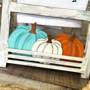 May include: Three wooden pumpkins in a white painted shelf. The pumpkins are painted teal, white, and orange.