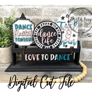 May include: A set of four wooden signs with a dance theme. The signs are painted black with white polka dots and feature the text "Dance recital tonight", "Dance life", "Twist & Spin", and "Love to Dance". The signs are displayed on a black wooden shelf with a wooden bead garland.