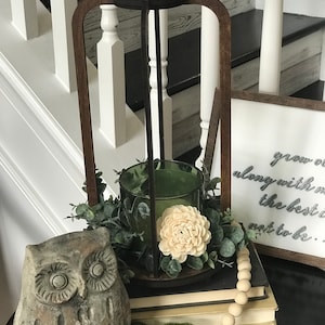 Op de afbeelding: Een decoratieve houten lantaarn in donkerbruin met een groene glazen kandelaar. De lantaarn is versierd met groen en een crèmekleurige bloem. Een uilbeeld en een snoer van houten kralen zijn ook aanwezig.