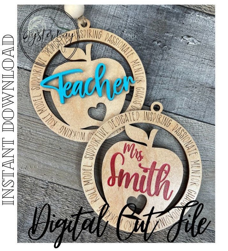 Puede incluir: Dos adornos de madera con forma de manzana con un dise&ntilde;o recortado. El adorno superior dice "Teacher" en letras azules y el adorno inferior dice "Mrs. Smith" en letras rojas. Ambos adornos tienen un c&iacute;rculo de texto alrededor de la manzana.