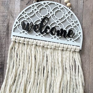 May include: White wooden welcome sign with a geometric pattern and black lettering. The sign is hanging from a string of wooden beads and has a fringe of white yarn.
