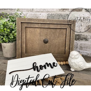以下が含まれることがあります： "home"のサインのデジタルカットファイル。サインは白い木でできており、黒い文字で書かれています。画像には、木製のフレーム、小さな鳥の置物、木製のビーズの紐も写っています。