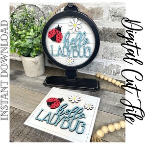 May include: A digital cut file for a "Hello Ladybug" sign. The sign features a red ladybug, white daisies, and the text "Hello Ladybug" in blue. The sign is designed to be cut from wood or other materials.