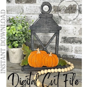May include: A gray wooden lantern with a black metal frame and a white X design. Two orange pumpkins are sitting inside the lantern. A string of wooden beads is in front of the lantern. The text "Digital Cut File" is written in black on a wooden surface.