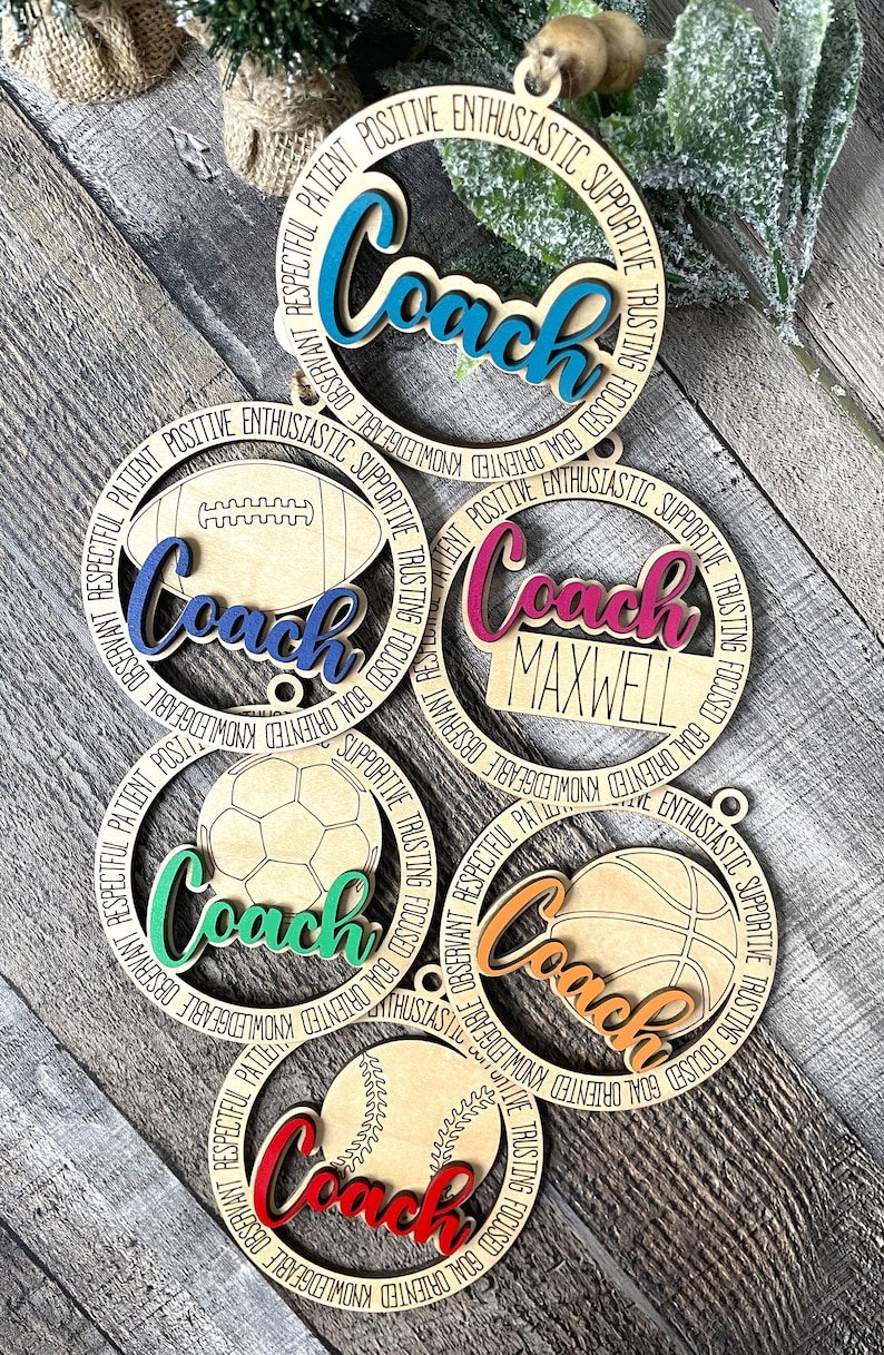 Peut inclure: Cinq ornements en bois avec diff&eacute;rents motifs de ballons de sport. Chaque ornement a un cercle de mots autour du bord, y compris "Coach", "Observant", "Respectueux", "Patient", "Positif", "Enthousiaste", "Soutien", "Confiant", "Concentr&eacute;", "Objectif", "Orient&eacute;" et "Connaissance".