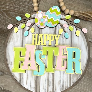 Puede incluir: Un letrero redondo de madera con un acabado blanqueado y un marco de madera marrón. El letrero dice "Feliz Pascua" en letras de colores con un contorno amarillo. Las letras están superpuestas y tienen un efecto 3D. Hay dos huevos de Pascua pastel con un esquema de colores verde, rosa y amarillo sobre el texto. Los huevos están decorados con lunares, zigzags y un círculo blanco. También hay ramas pastel verdes, rosas y azules con hojas alrededor de los huevos.