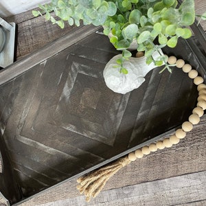 Op de afbeelding: Een zwarte houten dienblad met een chevron-patroon en een wit keramisch decoratief object. Het dienblad heeft twee handvatten en is versierd met een houten kralensnoer.