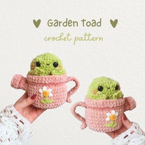 May include: Two plush crochet garden toads, each sitting inside a pink yarn watering can decorated with a white daisy. The text above reads Garden toad crochet pattern with two small heart icons on either side of the title text.