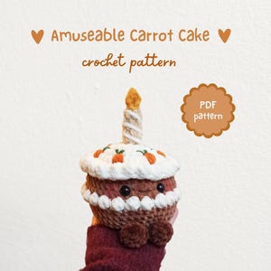 May include: A crocheted carrot cake with a candle, featuring white frosting with orange carrot decorations. The cake has brown and white details, a smiling face and small brown feet. Text reads "Amuseable Carrot Cake crochet pattern".