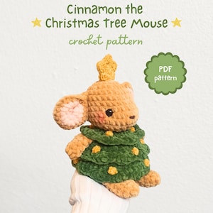 Peut inclure: Une souris au crochet vêtue d'un costume de sapin de Noël vert avec des accents jaunes. La souris a une couronne en forme d'étoile et des oreilles intérieures roses. Le texte sur l'image indique "Cinnamon the Christmas Tree Mouse crochet pattern" et "PDF pattern."