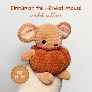 Peut inclure: Une souris crochetée aux tons bruns et orangés, avec des intérieurs d'oreilles roses et des yeux noirs. Le texte "Cinnamon the Harvest Mouse crochet pattern" est affiché. Un badge "PDF pattern" est également visible.