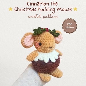 May include: A crocheted mouse toy in the shape of a Christmas pudding, with a brown body, white collar, and green holly accents. The text "Cinnamon the Christmas Pudding Mouse crochet pattern" is displayed above the toy.